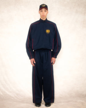Model wearing the navy and red piped retro track pant with a matching track jacket with crest patch and a cap