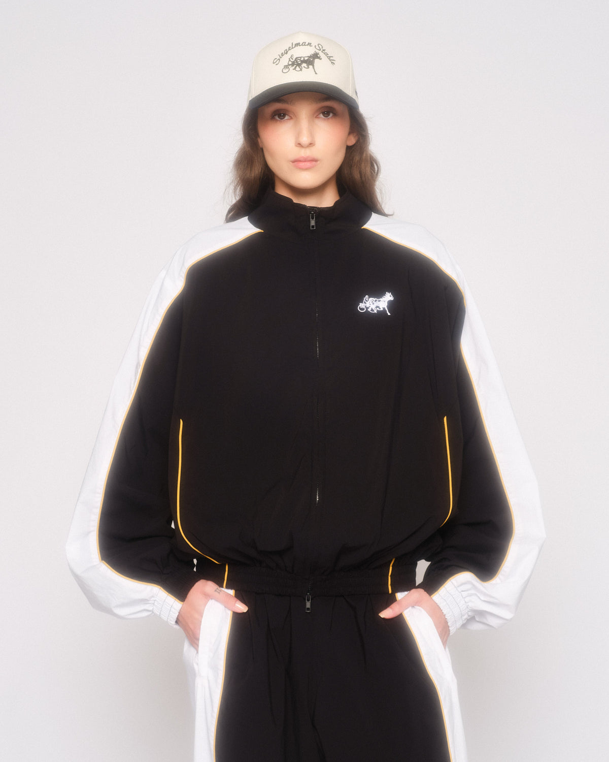 Model wearing the black and white track jacket with yellow piping, matching track pants and a cream cap