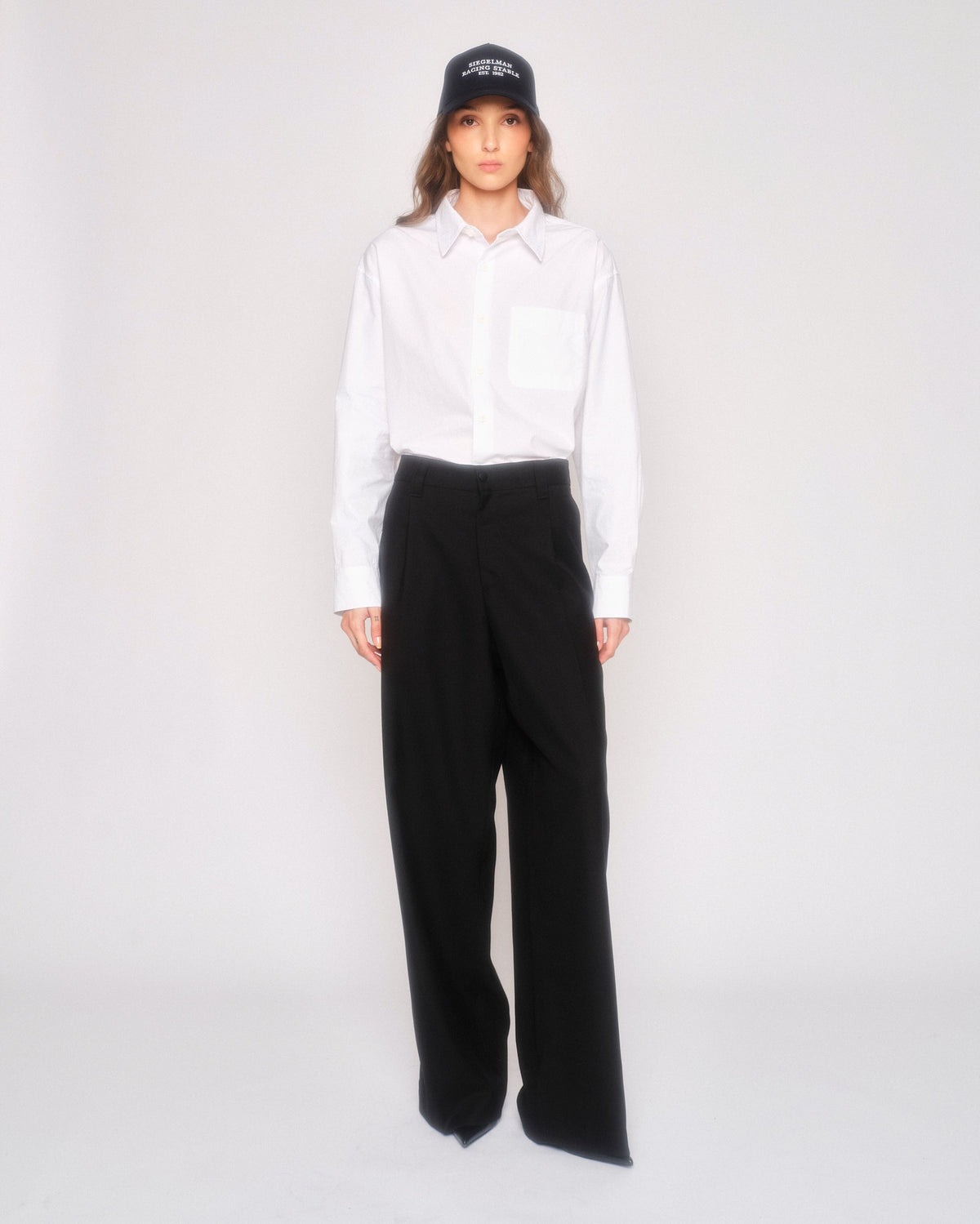 Model wearing the black pleated wide leg pants with a white button-up shirt and navy cap