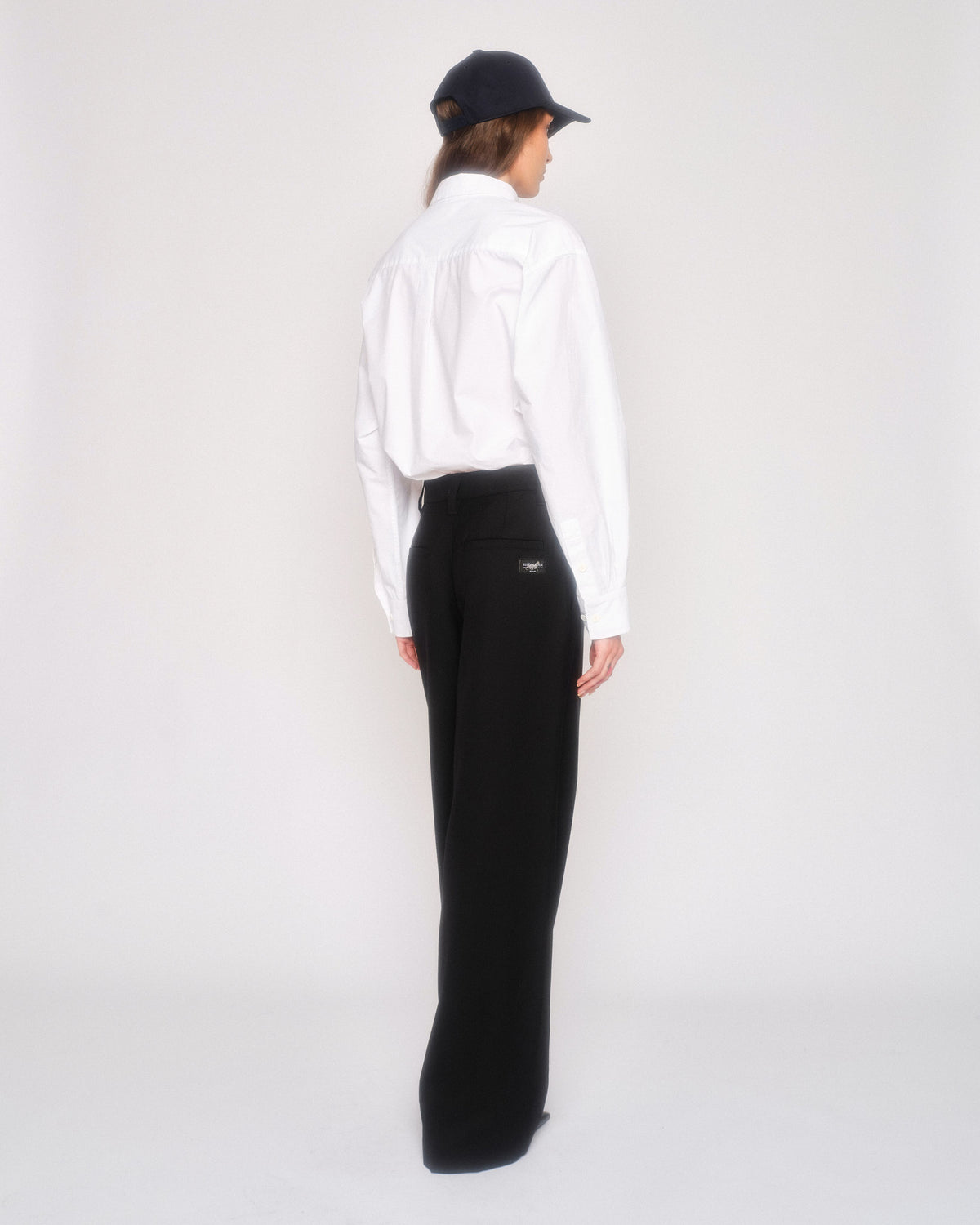 Back three-quarter view of a model in the black pleated pants, white shirt and navy cap