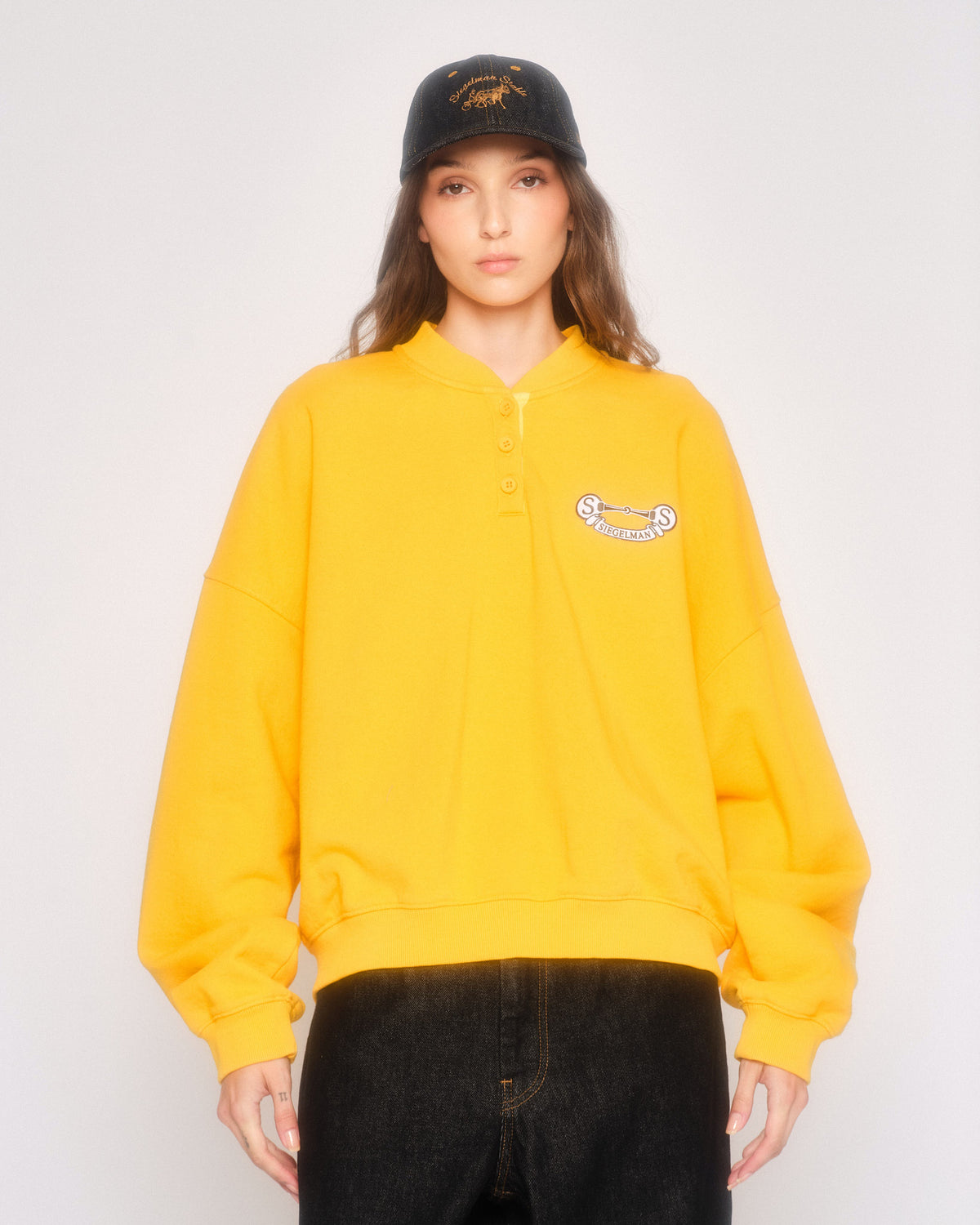 Model wearing the yellow henley sweatshirt with crest patch, a black cap and black jeans