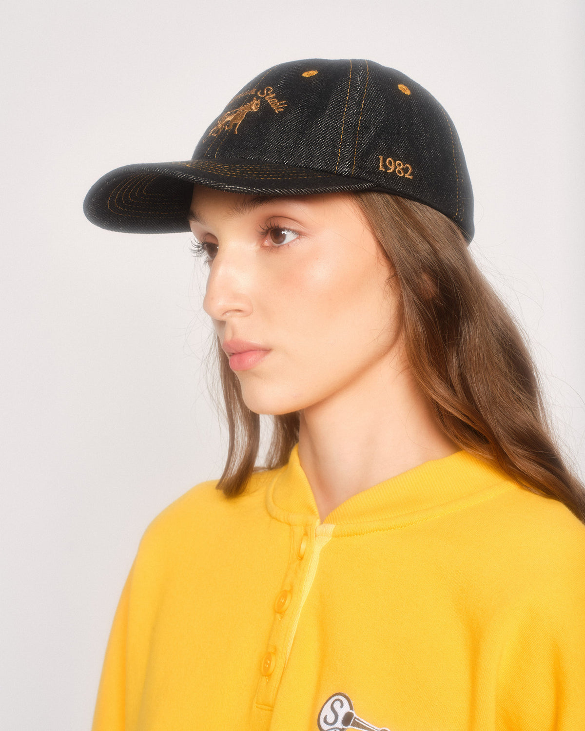 Model in three-quarter view wearing the black denim dad hat with gold stitching and gold 1982 side embroidery
