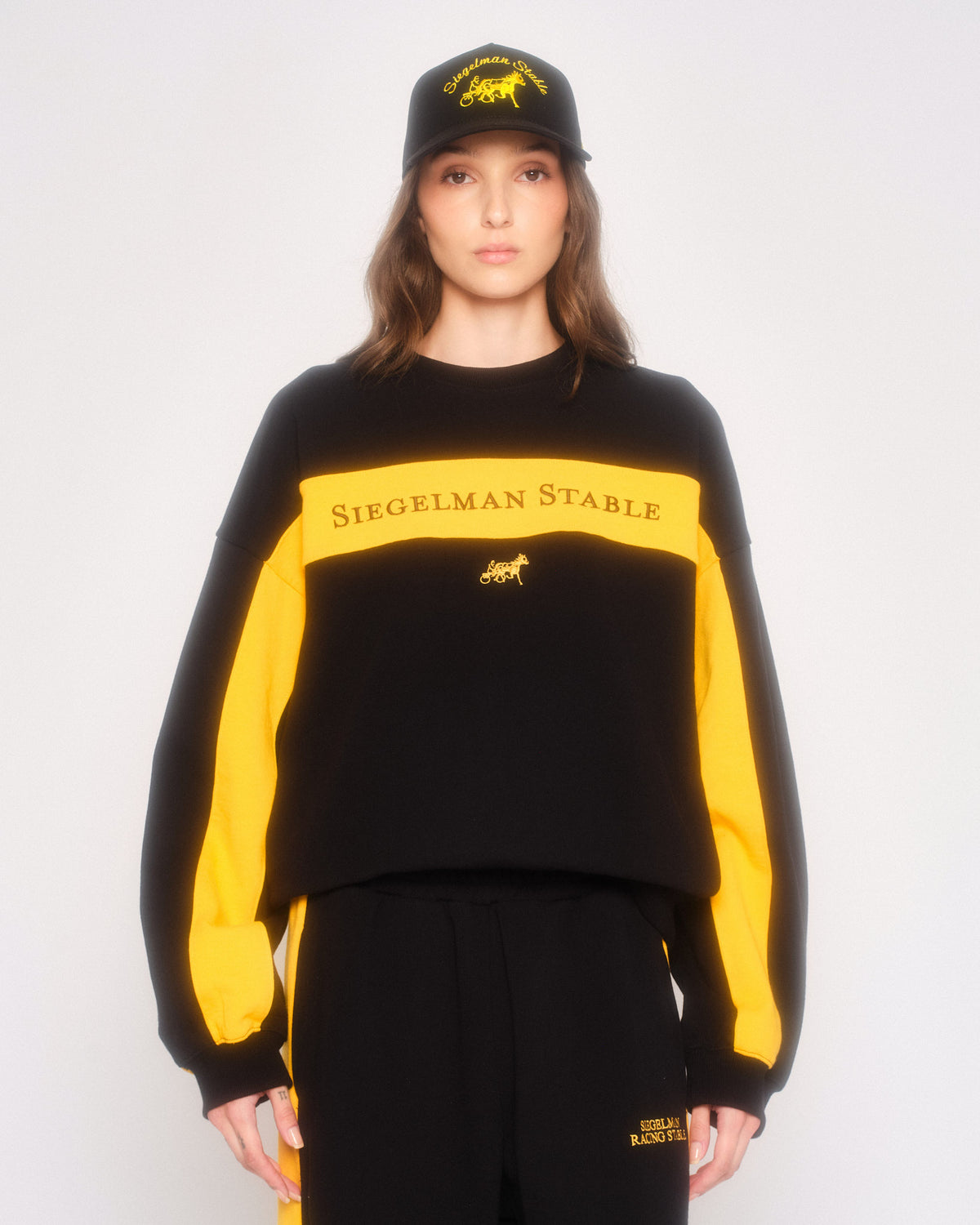 Model wearing the black and yellow Siegelman Stable retro crewneck with matching sweatpants and black cap