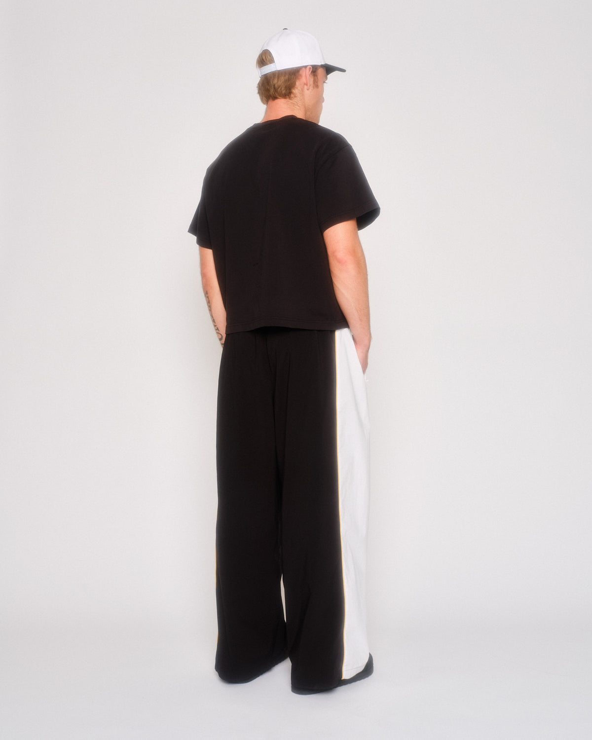 Back three-quarter view of a male model in the black wide leg track pants and white cap