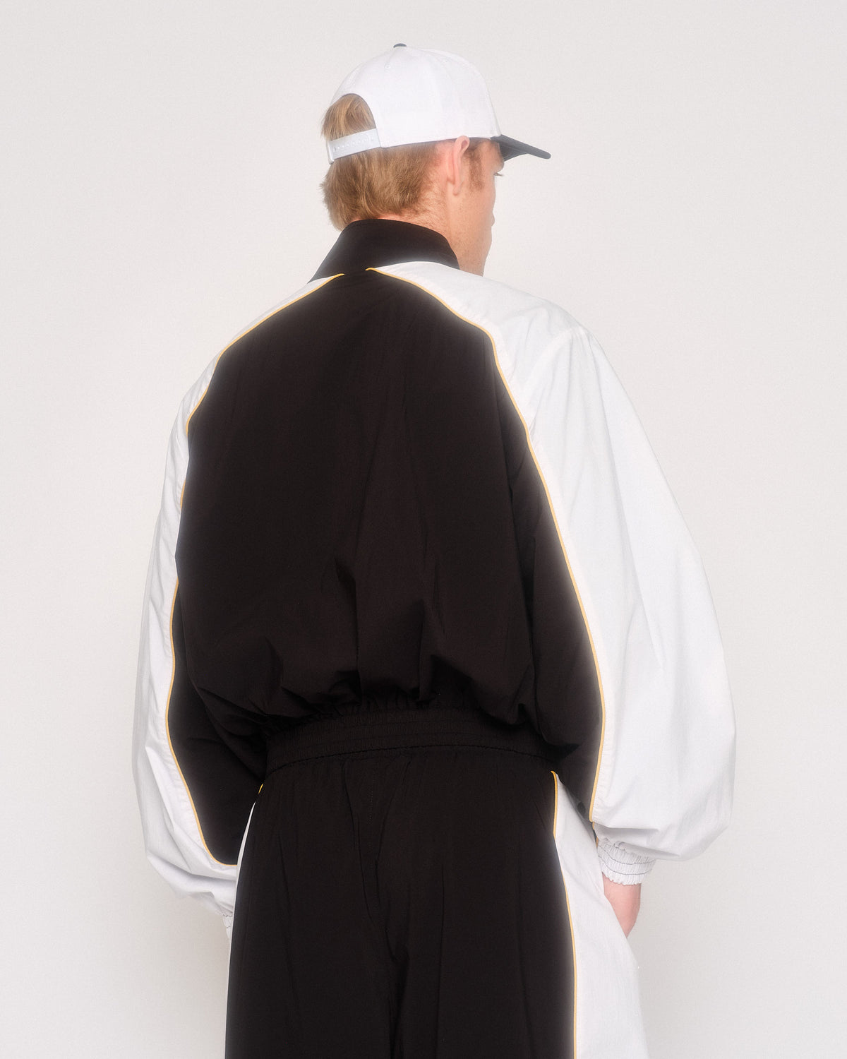 Back view of a model in the black track jacket with white raglan sleeves and a white cap