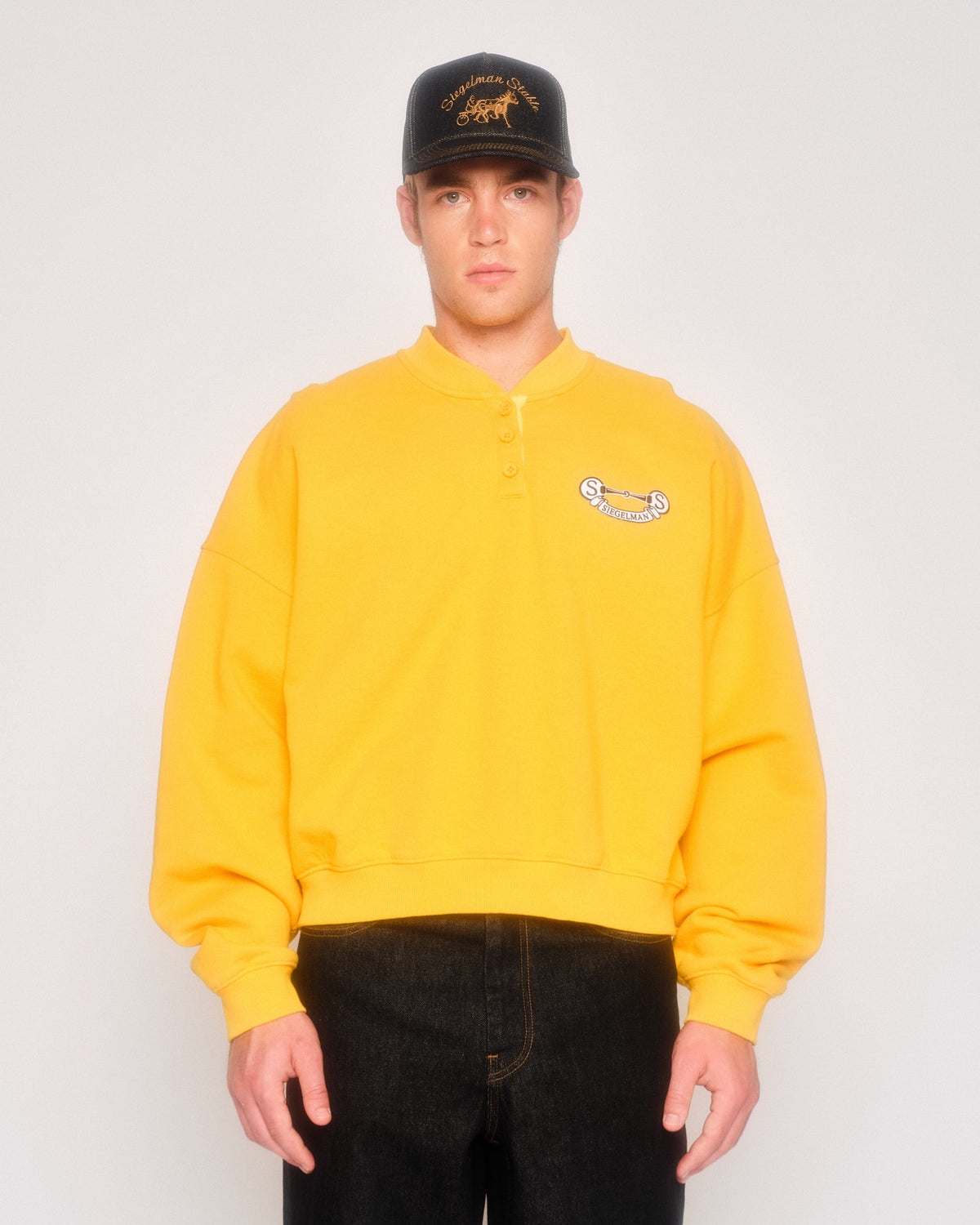 Male model wearing the yellow henley sweatshirt with crest patch, a black cap and black jeans