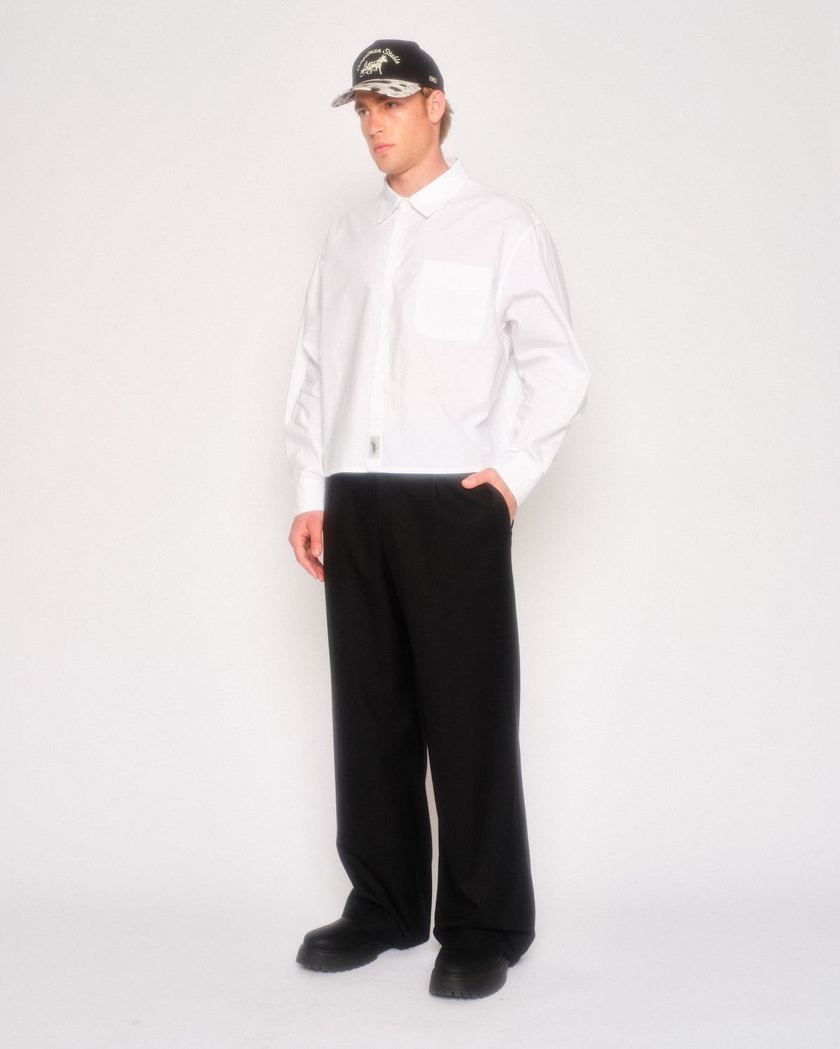 Male model in three-quarter view wearing the black pleated trousers, white shirt and black cap