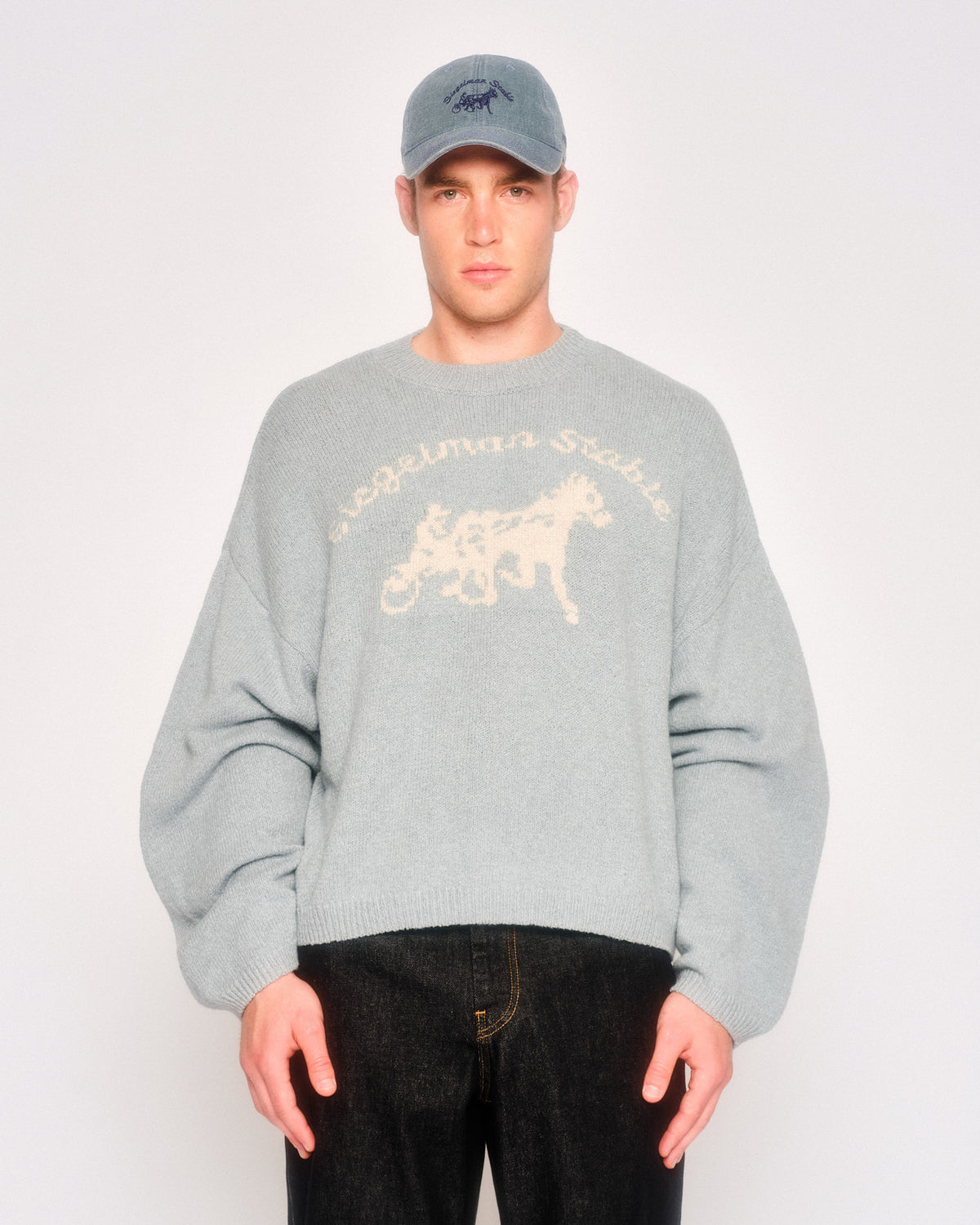 Model wearing the dusty blue Siegelman Stable logo knit sweater with a gray-blue cap and dark jeans