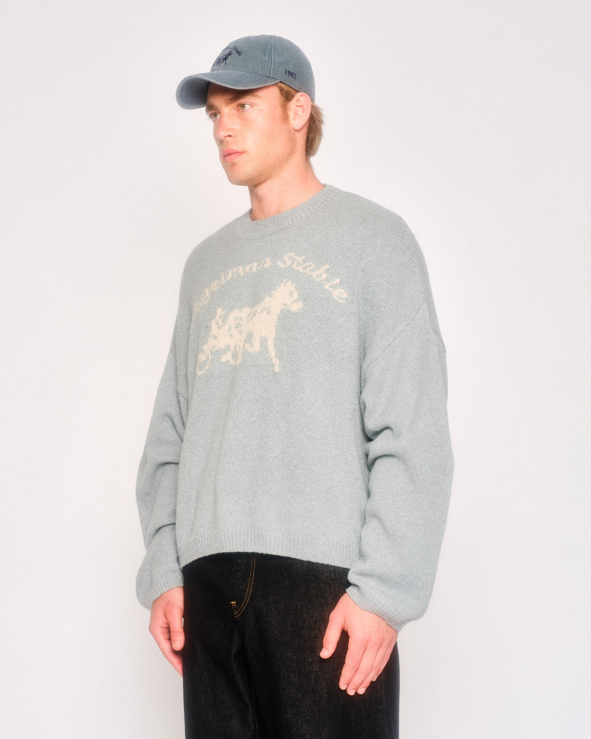 Model in three-quarter view wearing the dusty blue logo knit with cream horse and sulky graphic and a gray-blue cap
