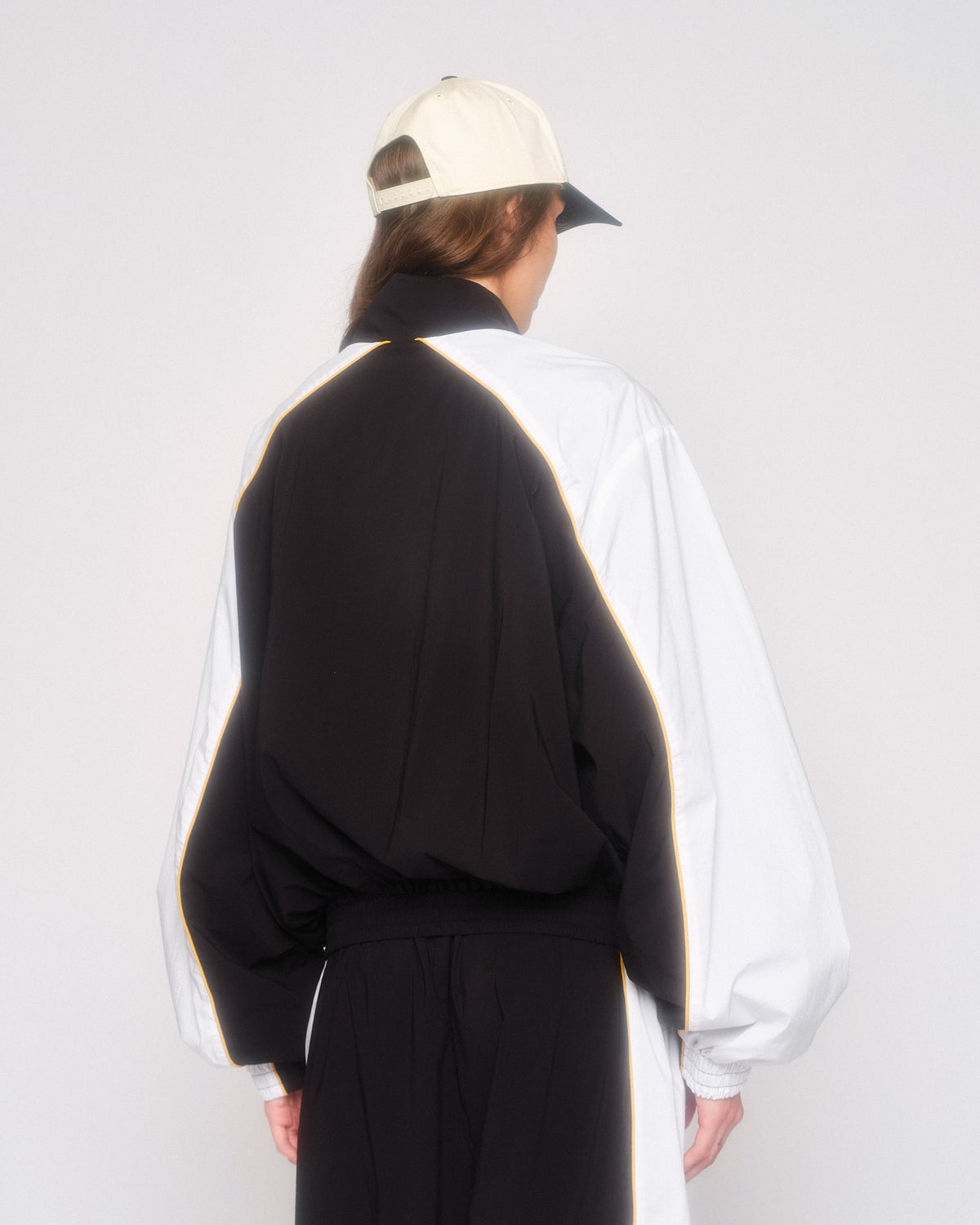 Back three-quarter view of a model in the black and white track jacket and cream cap