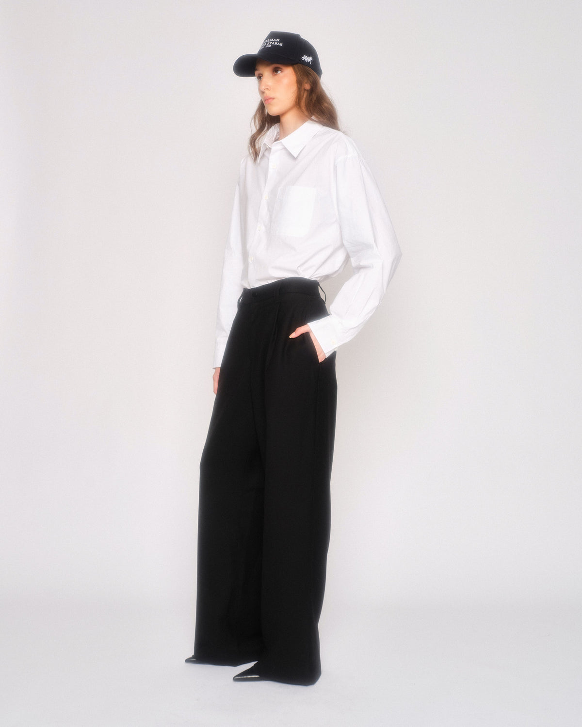 Model in profile wearing the black pleated trousers with a hand in pocket and a white shirt