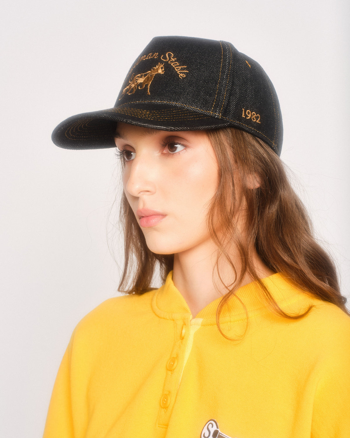 Model in profile wearing the black denim hat with gold stitching and gold 1982 side embroidery