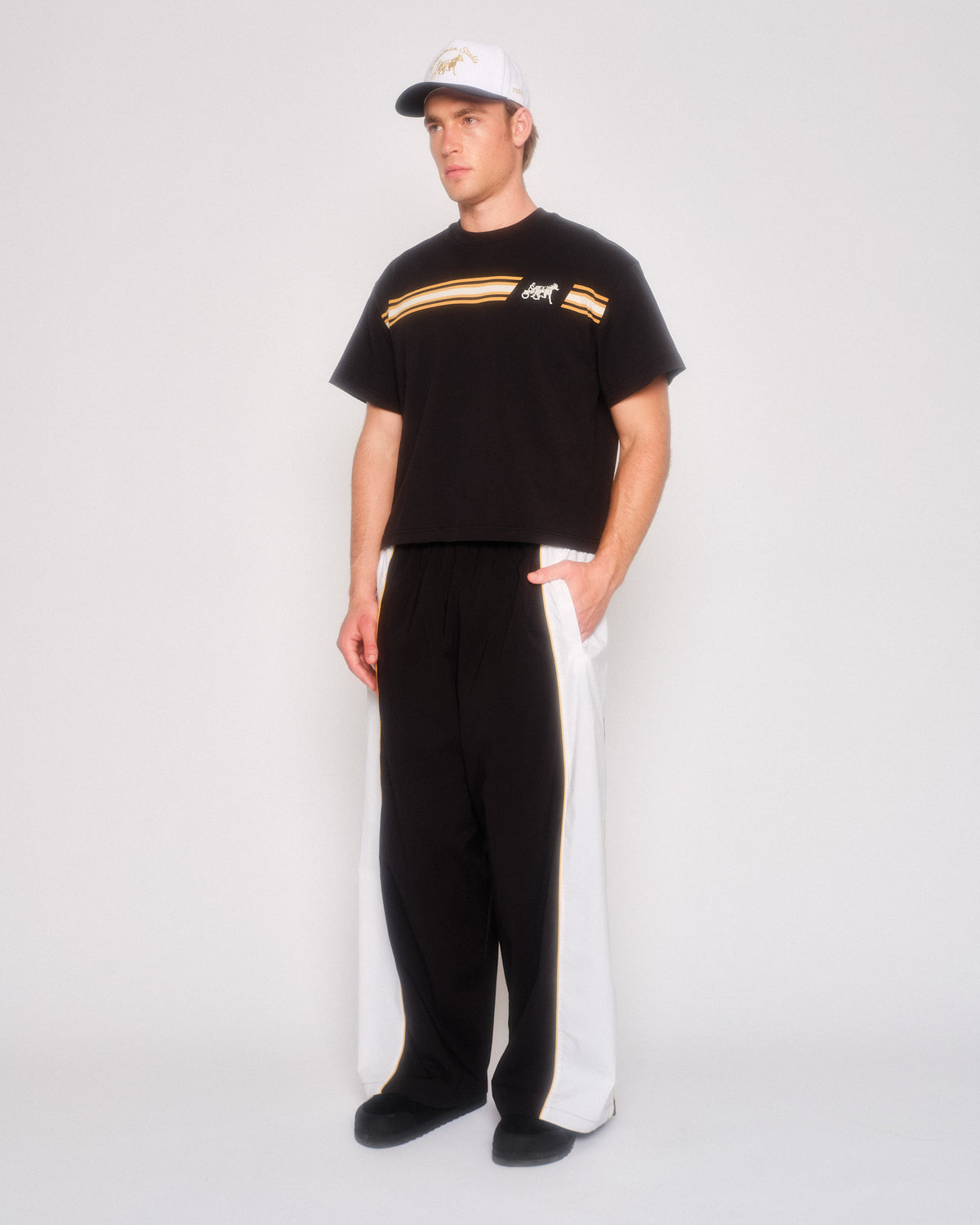 Male model in three-quarter view wearing the black and white track pants, black tee and white cap