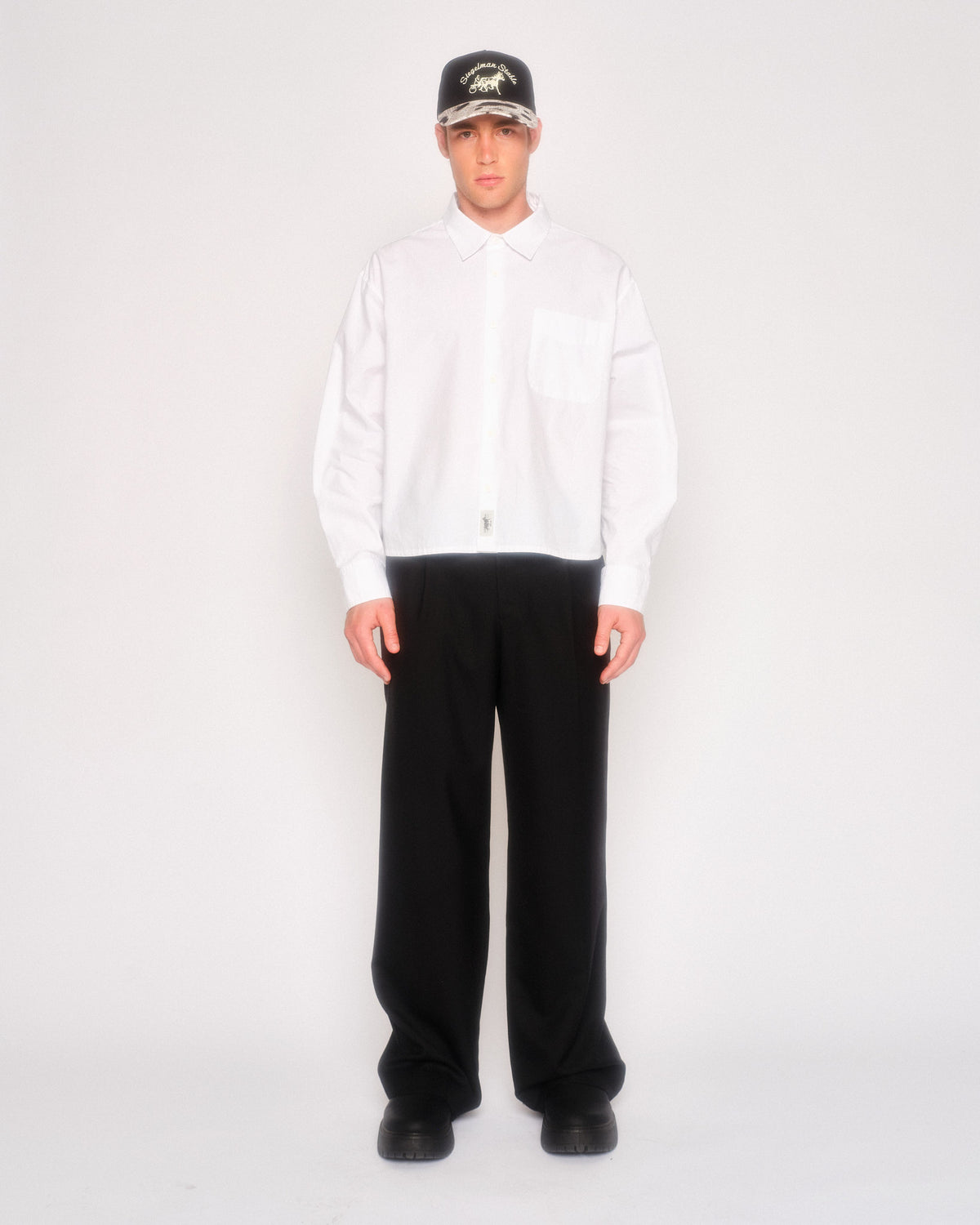 Male model wearing the black pleated pants with a white button-up shirt and black cap